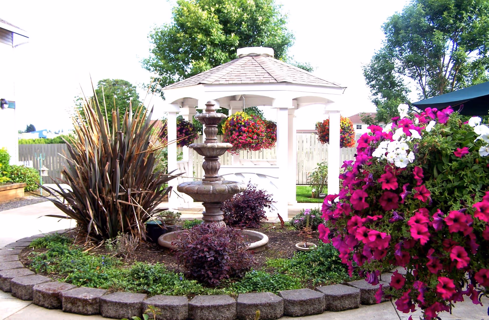 Outdoor garden area with a stone fountain in the center surrounded by various plants and flowers. In the background, there is a white gazebo with hanging flower baskets and trees around the space.