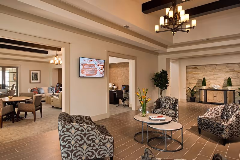 A spacious and well-lit common area in a senior living facility featuring patterned armchairs around a round coffee table with a vase of yellow flowers. In the background, there is a dining area with a round table and chairs, a wall-mounted TV, and an office space with a desk and chair. The room has beige walls, wooden flooring, and decorative ceiling beams with modern chandeliers.