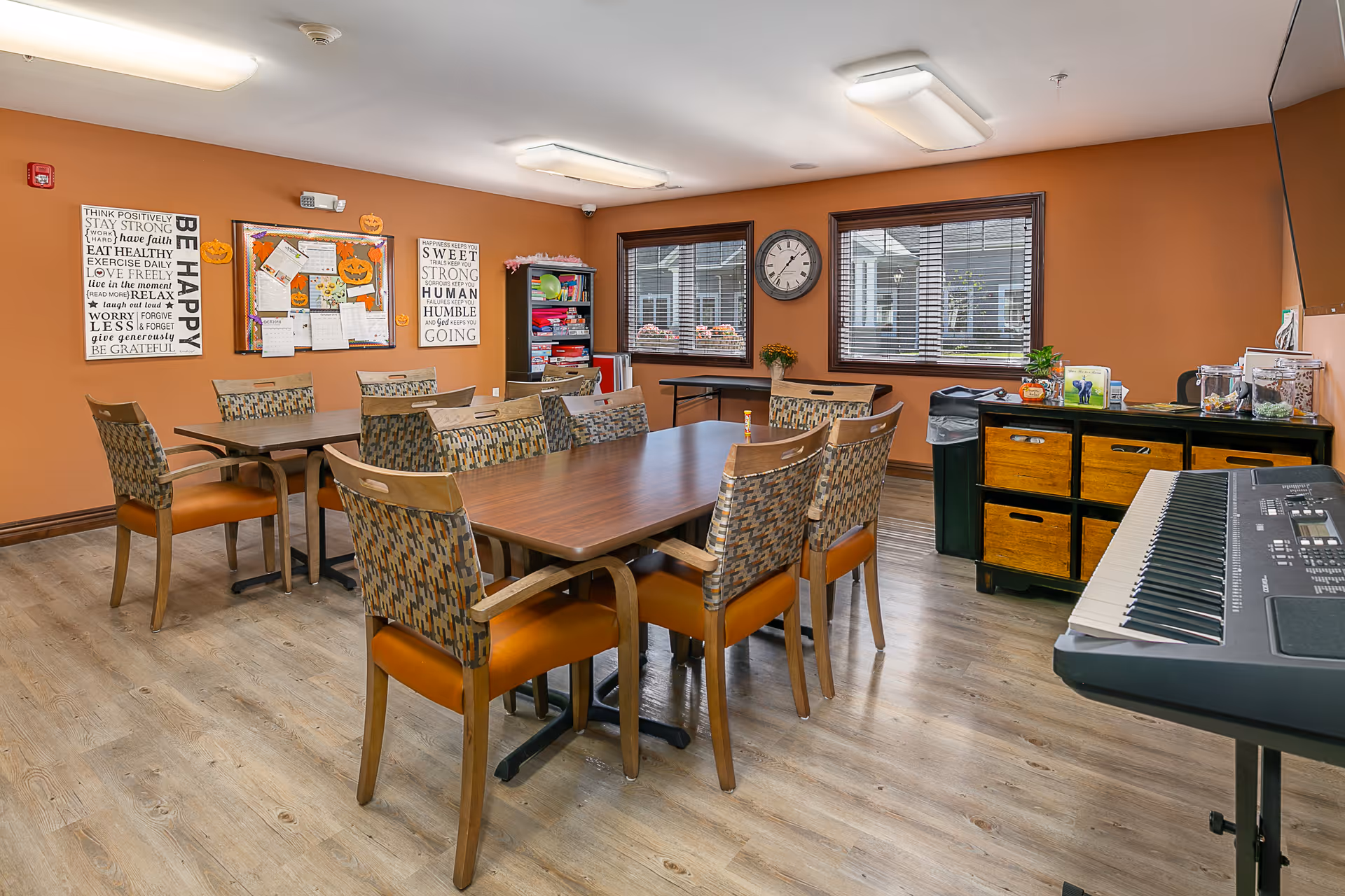 A well-lit common room with two wooden tables surrounded by patterned chairs with orange cushions. The walls are painted a warm brown color and decorated with motivational posters and a bulletin board with various papers and Halloween decorations. There are two windows with blinds, a clock on the wall, a black cabinet with wooden drawers, and a keyboard on a stand in the foreground.