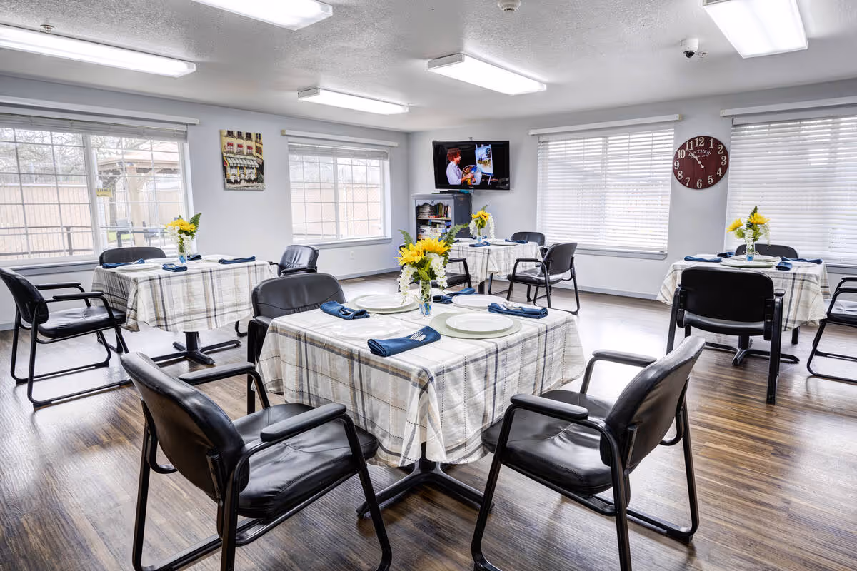 A bright dining room with four tables covered in plaid tablecloths, each set with plates, blue napkins, and vases of yellow flowers. Black chairs surround the tables. Large windows with white blinds let in natural light. A wall clock and a TV mounted on the wall are visible in the background.