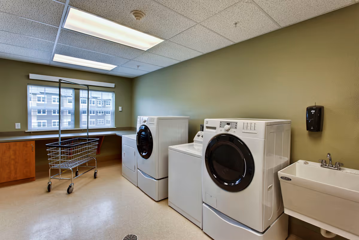 Community laundry room with front-loading washer and dryer, utility sink, folding counter, and a rolling laundry cart.