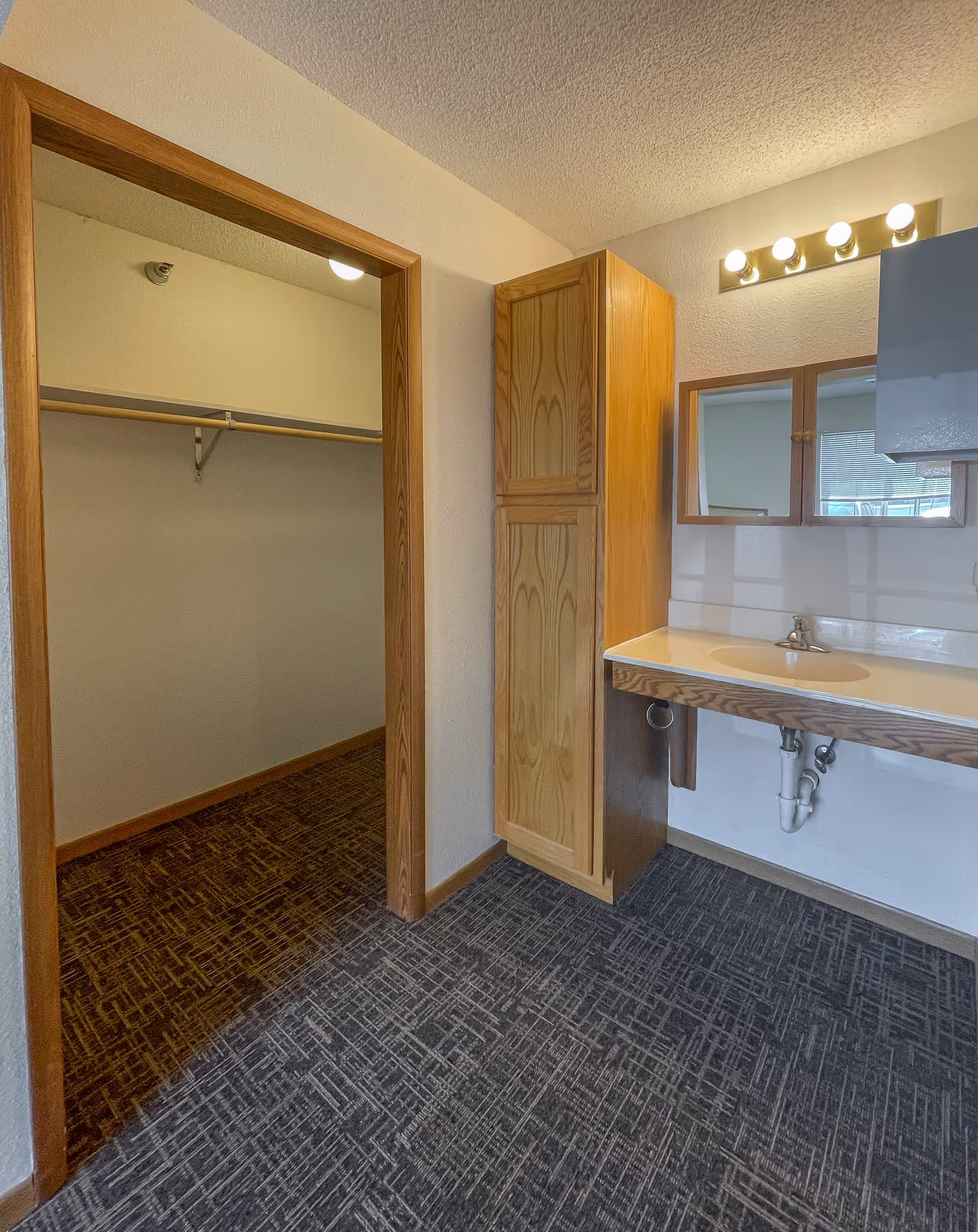 Interior view of a room in Park Lane Estates featuring a wooden framed mirror above a sink with a countertop, a tall wooden cabinet, and an open doorway leading to a walk-in closet with a hanging rod and shelf. The room has patterned carpet flooring and white walls.