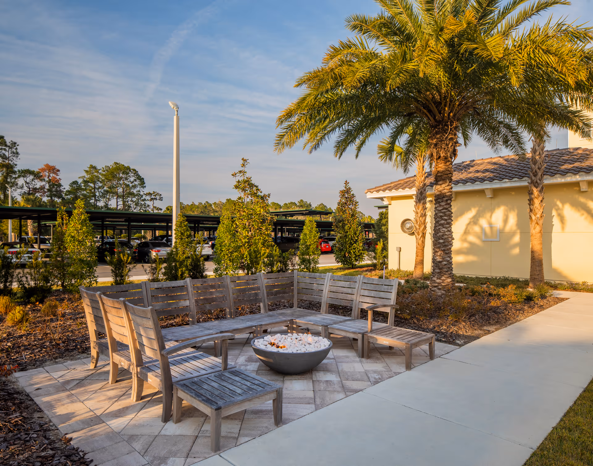 Outdoor seating area with wooden benches arranged in a semi-circle around a fire pit filled with white stones, surrounded by palm trees and landscaping. In the background, there is a parking area with covered carports and several parked cars under a partly cloudy sky.