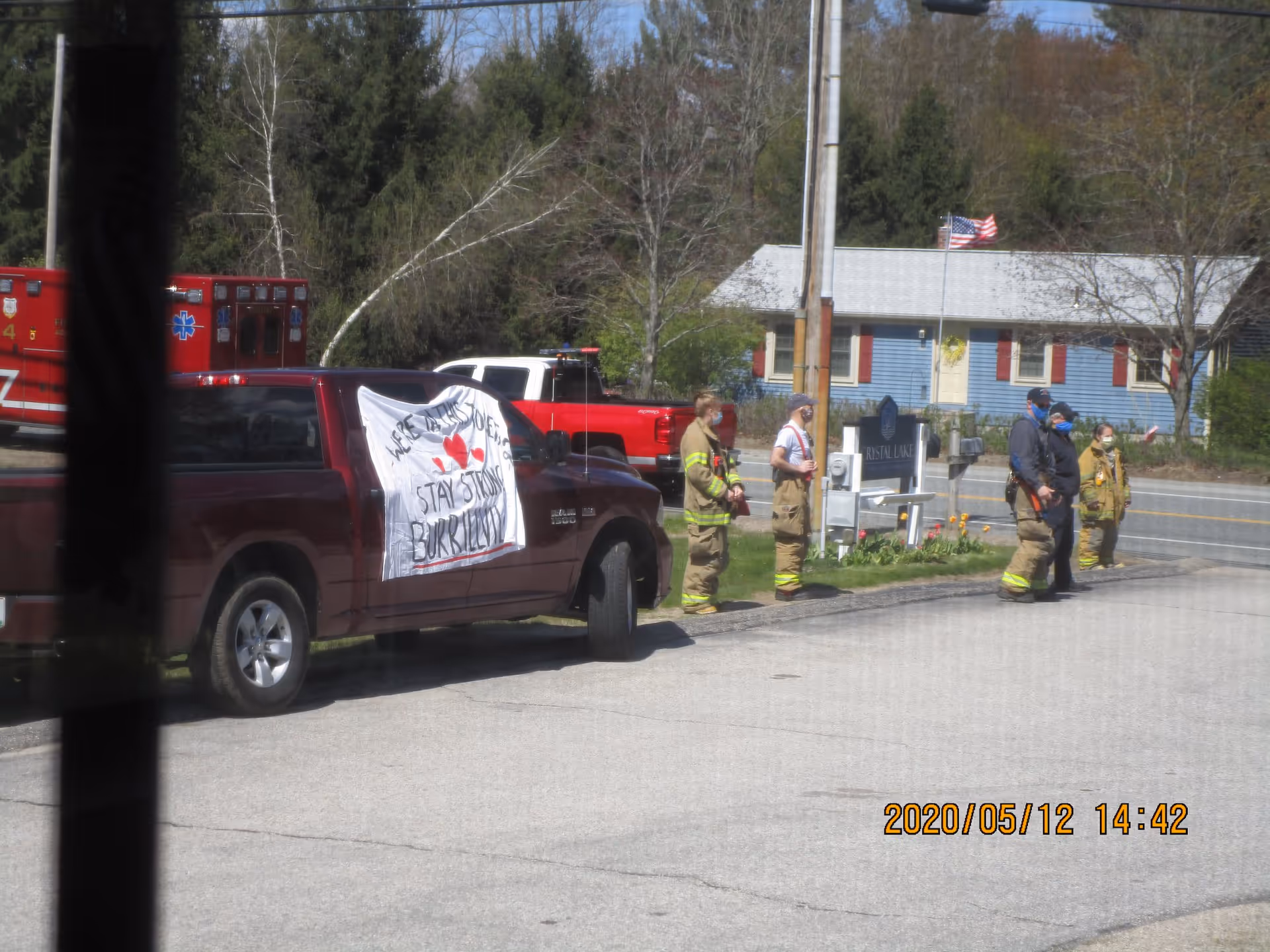 A group of firefighters and emergency responders stand outside near a red pickup truck with a white banner that reads 'We're all in this together, stay strong Burkeville' next to a sign for Crystal Lake Rehabilitation and Care Center. The scene is set on a sunny day with trees and a blue house in the background.