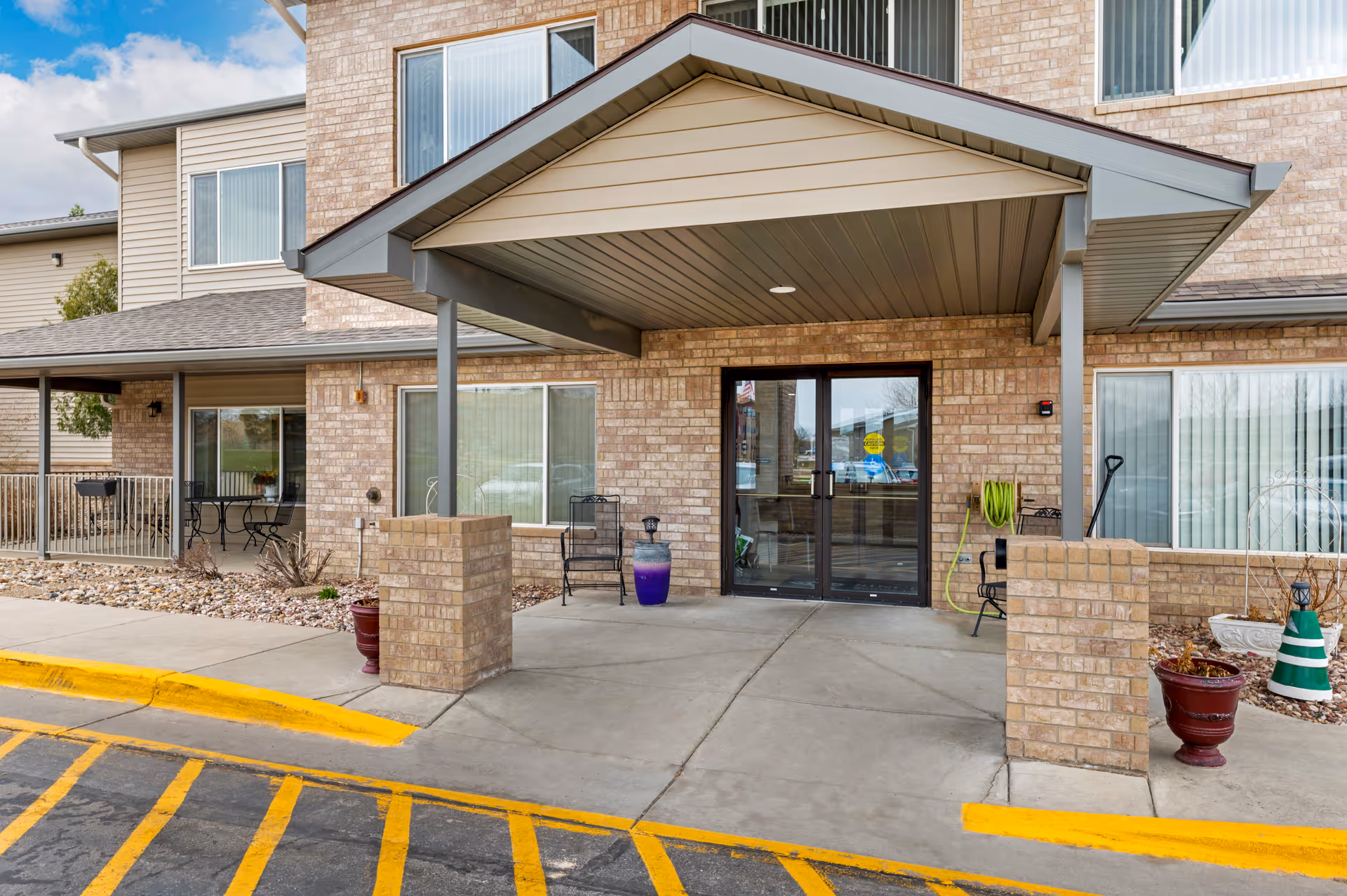 Entrance of a senior living facility with a covered porch supported by two pillars, glass double doors, brick exterior walls, and windows with vertical blinds. There are potted plants and outdoor chairs near the entrance, and a yellow-striped parking area in front.