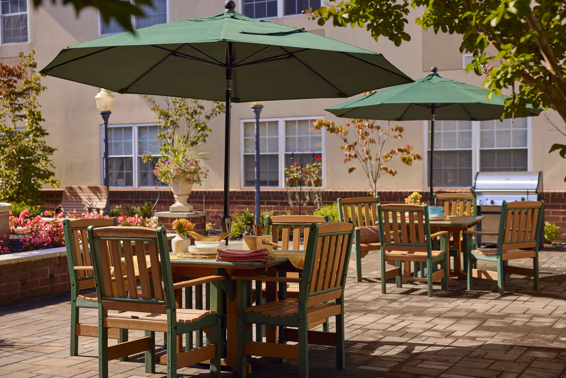 Outdoor patio area with two wooden tables and green umbrellas providing shade. Each table is surrounded by wooden chairs with green frames. The patio is paved with bricks and decorated with potted plants and flowers. A stainless steel grill is visible near the building wall in the background.