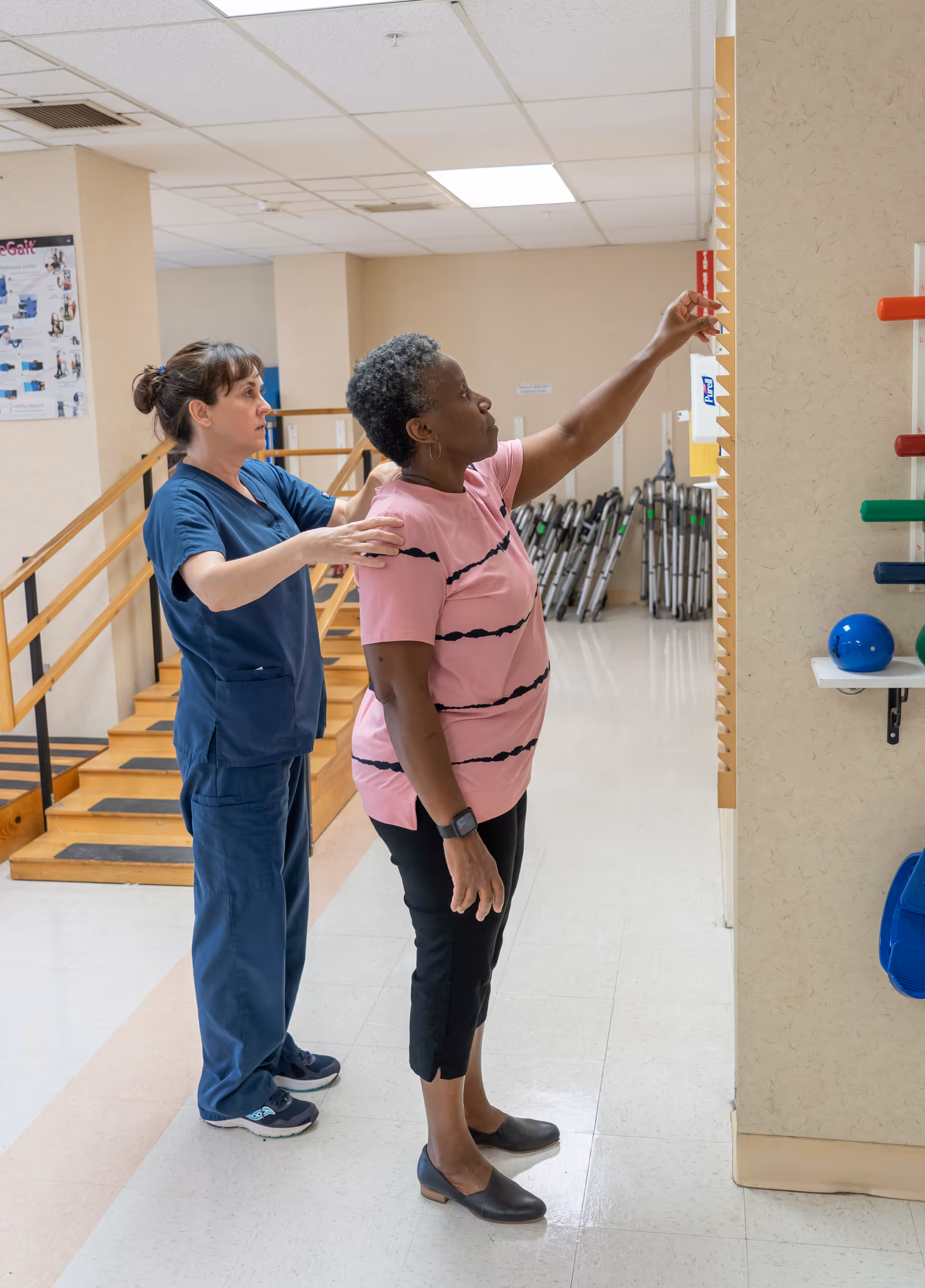 A healthcare worker in blue scrubs assists an older woman in a pink shirt and black pants as she reaches up to a height measurement chart in a rehabilitation or physical therapy room with exercise equipment and parallel bars in the background.