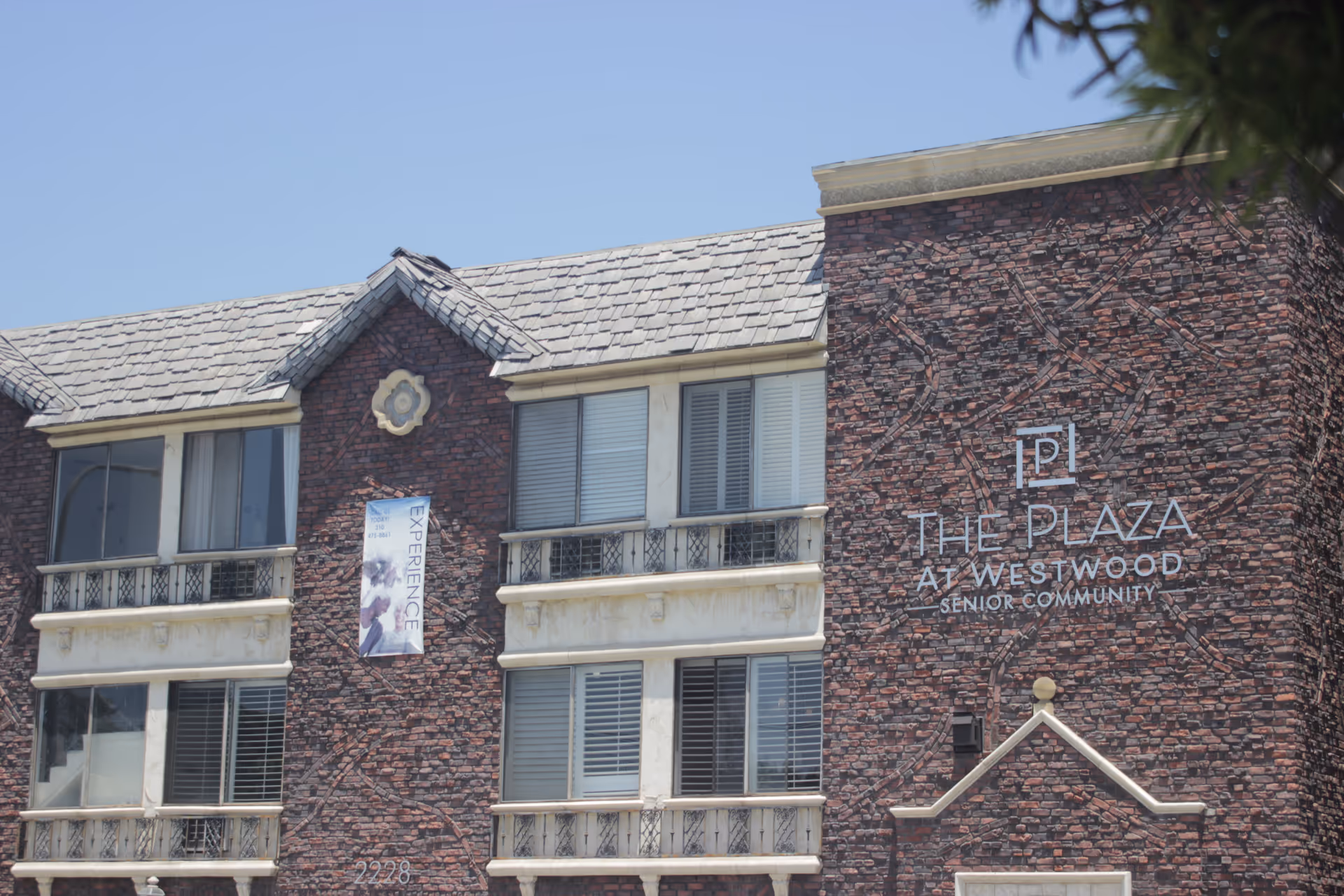 Exterior view of The Plaza At Westwood Senior Community building with brick facade, multiple windows, and a banner that says EXPERIENCE.