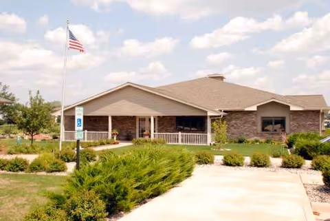 Single-story senior facility with stone facade, covered entrance, manicured shrubs, and flag.