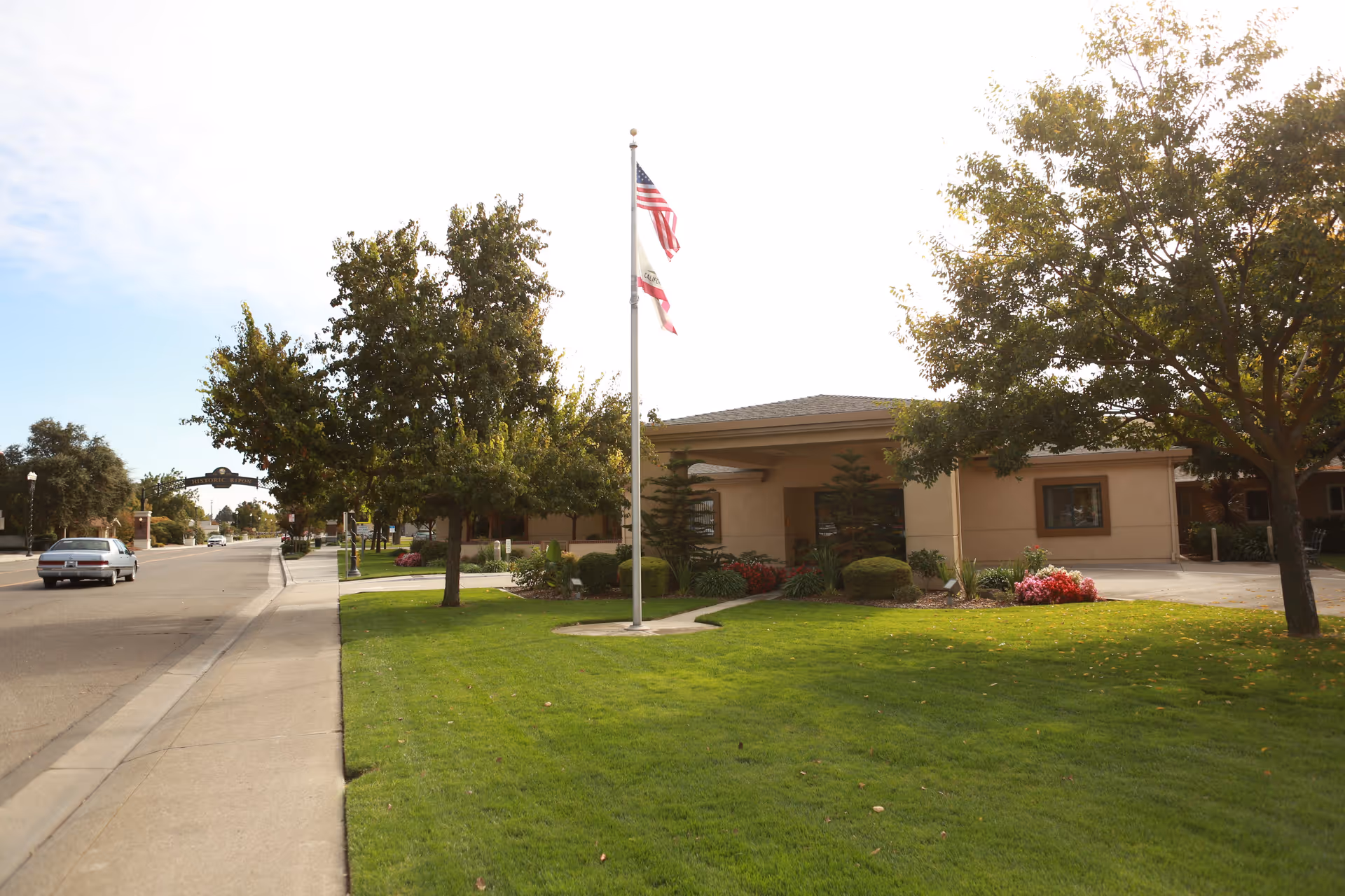Exterior view of Bethany Home Society San Joaquin County building with a well-maintained lawn, trees, and two flagpoles displaying the American and California state flags. A sidewalk and street run alongside the property, with a car driving on the road.