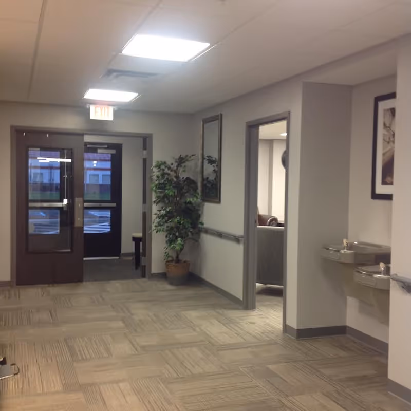 Interior hallway of Hyde Park Assisted Living Facility with beige carpeted floor, light gray walls, two drinking fountains mounted on the right wall, a potted plant and a mirror on the left wall, and double glass doors leading outside at the end of the hallway. There is an open doorway on the right showing a glimpse of a room with chairs.