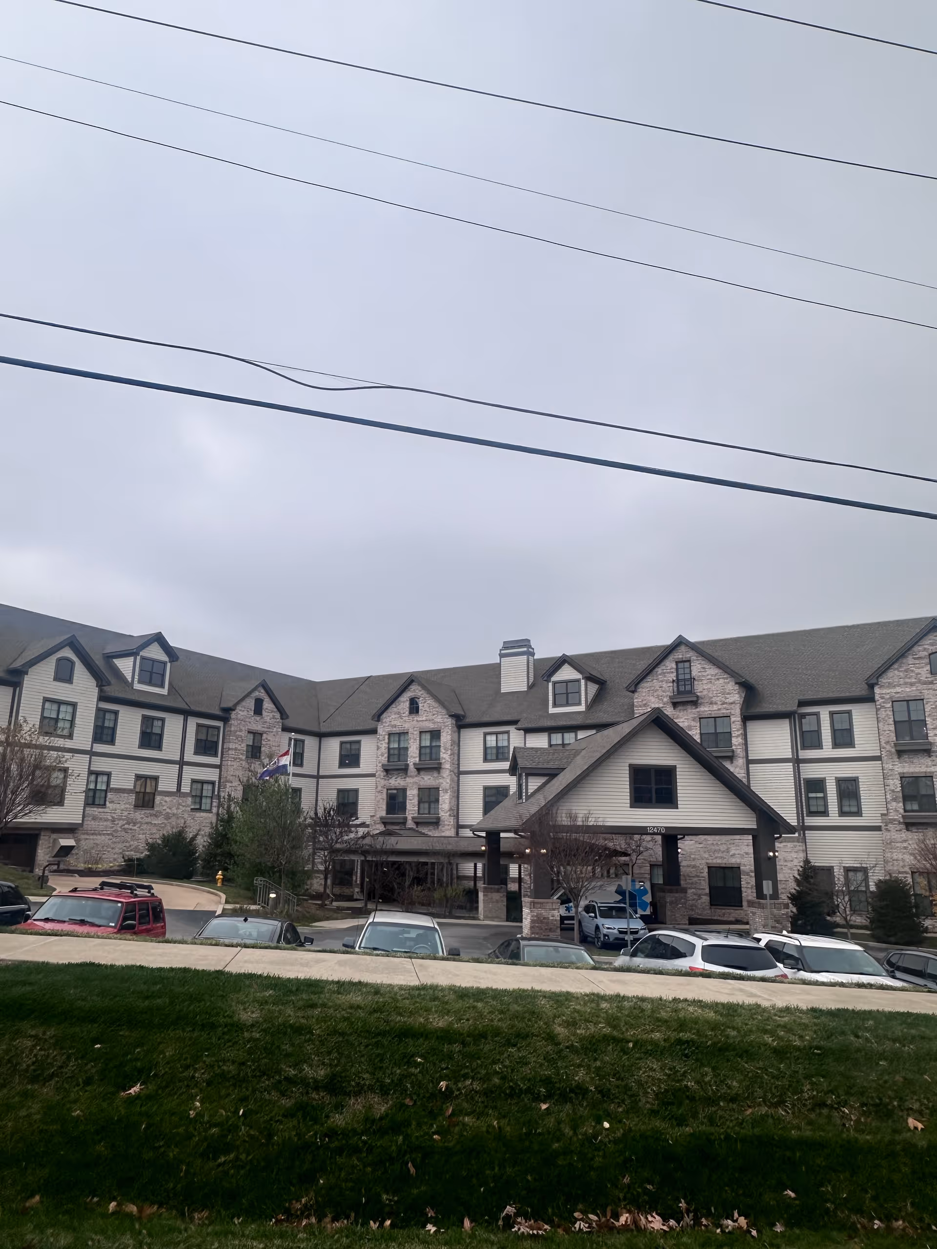 Exterior view of The Grande At Laumeier Park, a multi-story senior living facility with a combination of brick and siding facade, multiple windows, and a covered entrance. Several cars are parked in front of the building, and there is a grassy area in the foreground under an overcast sky.