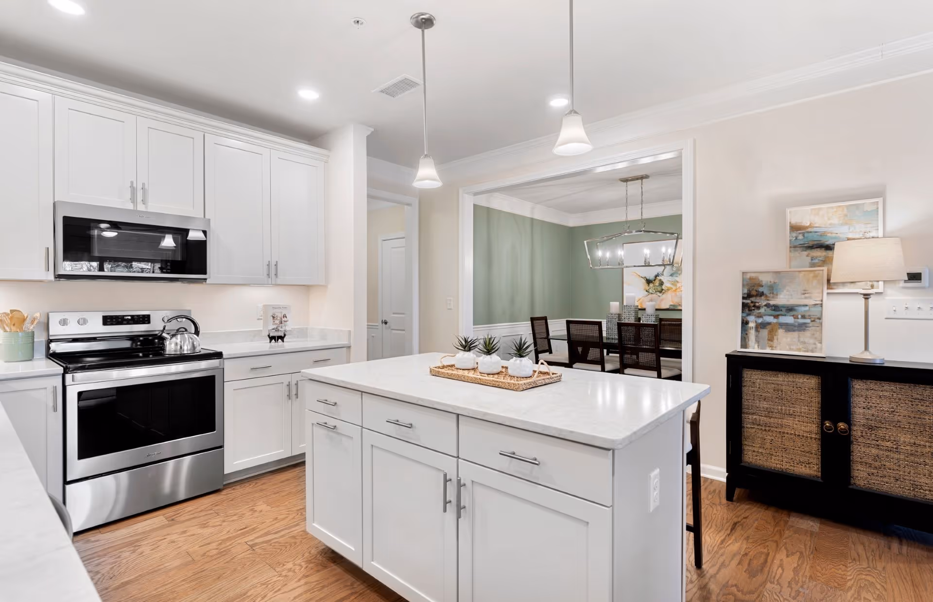 Modern kitchen with white cabinetry, stainless steel stove and microwave, a central island with small potted plants on a tray, hardwood flooring, and pendant lights overhead. In the background, a dining room with a green accent wall, a rectangular chandelier, and a dining table with chairs is visible. There is also a black sideboard with woven cabinet doors, a table lamp, and framed artwork on the right side.