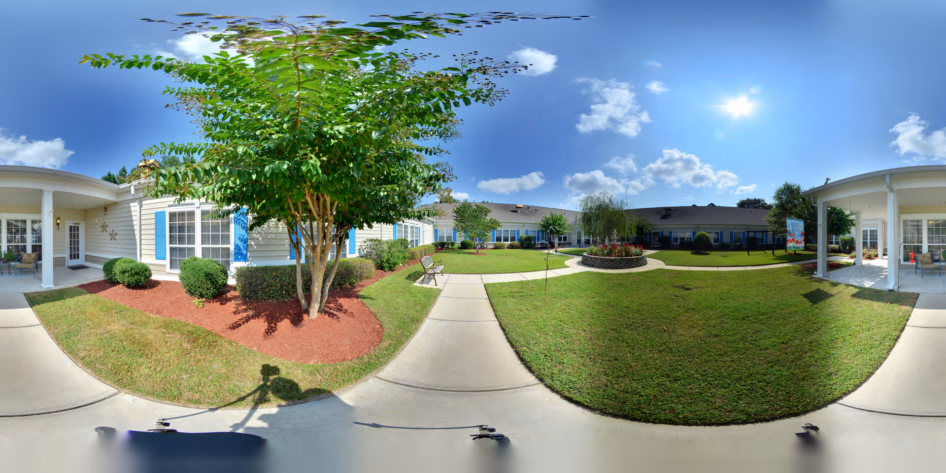 A sunny outdoor courtyard at The Legacy of Orangeburg featuring a well-maintained lawn, a circular flower bed with colorful plants, several benches along the paved walkways, and a few small trees and shrubs. The surrounding building has light-colored siding with blue shutters and covered patio areas with seating.