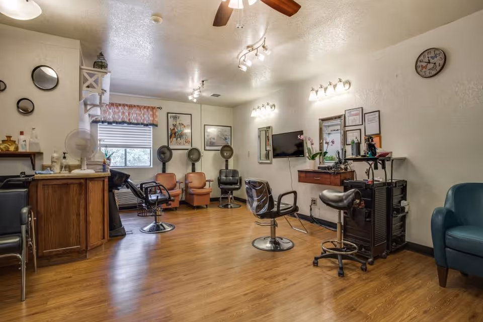 Interior view of a hair salon area in a senior living facility with wooden flooring, salon chairs, hair dryers, a wall-mounted TV, mirrors, and various hair care products on shelves and carts. The room is well-lit with ceiling lights and has a window with a patterned valance.