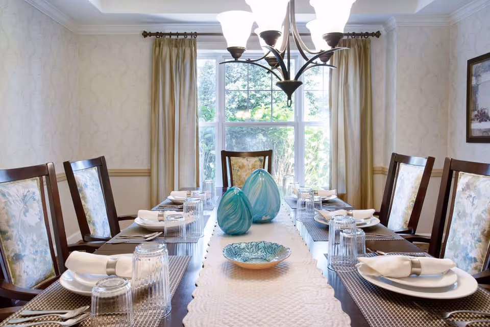 A dining room with a long wooden table set for six people. Each place setting includes a white plate, a folded white napkin with a napkin ring, and an upside-down clear drinking glass. The table has a white textured runner with two blue decorative vases and a blue patterned bowl in the center. The room has beige patterned wallpaper, a large window with beige curtains, and a chandelier hanging above the table.