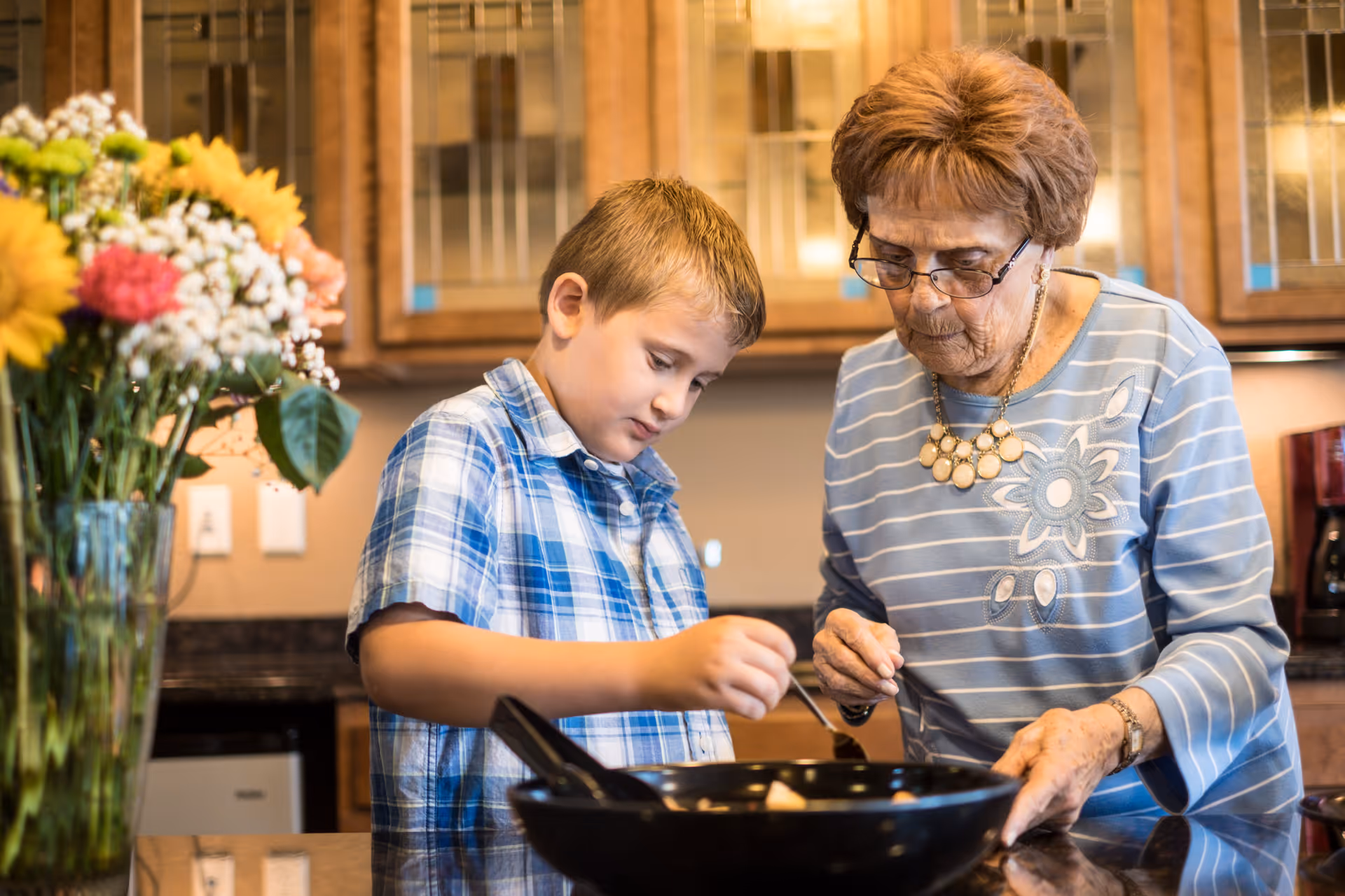 An elderly woman and a young boy cooking together in a kitchen. The boy is stirring food in a pan while the woman watches closely. There is a vase with colorful flowers on the counter nearby.