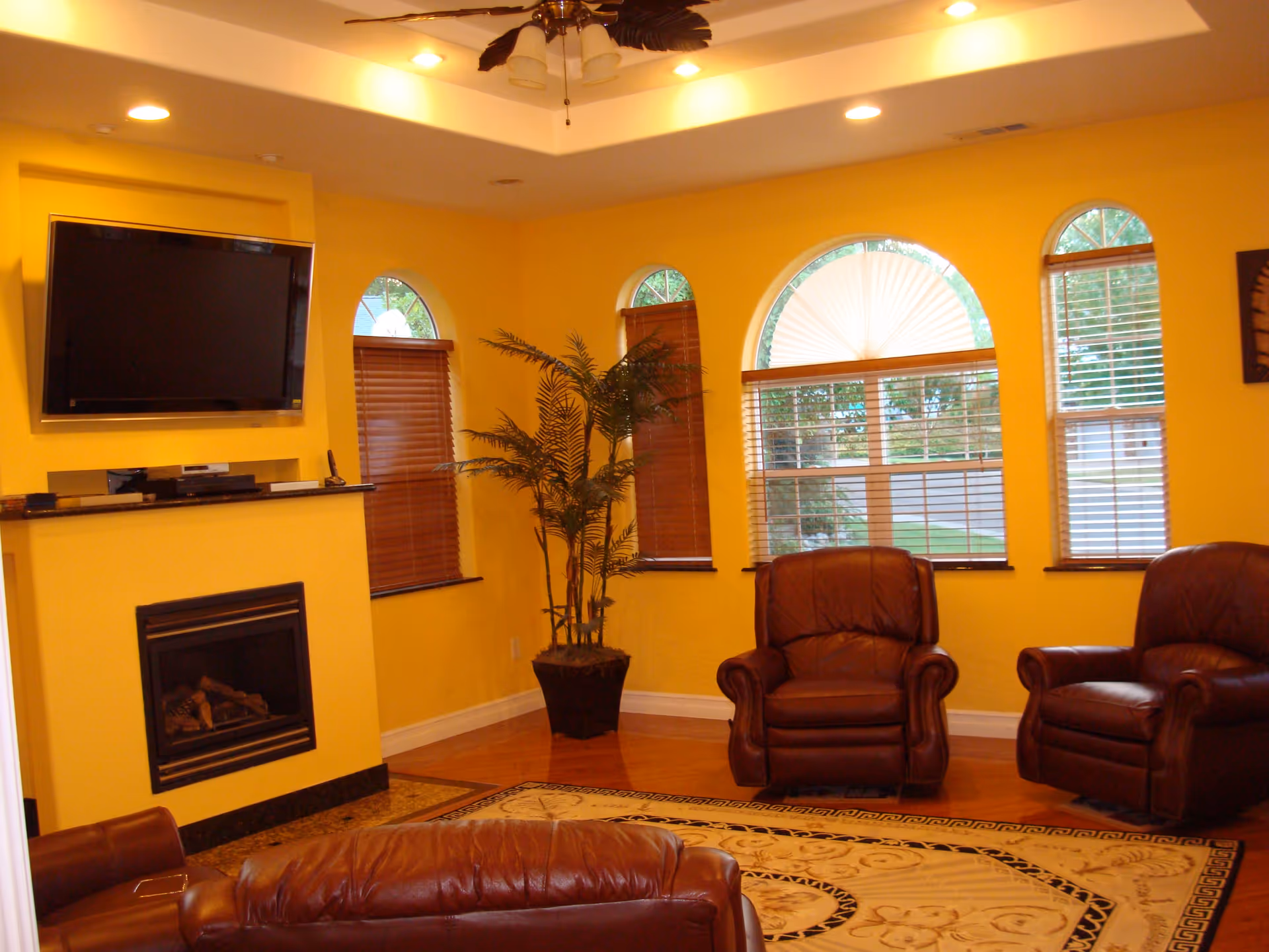 A cozy living room with bright yellow walls, three arched windows with wooden blinds, a mounted flat-screen TV above a fireplace, a potted plant in the corner, and three brown leather armchairs arranged around a patterned area rug.
