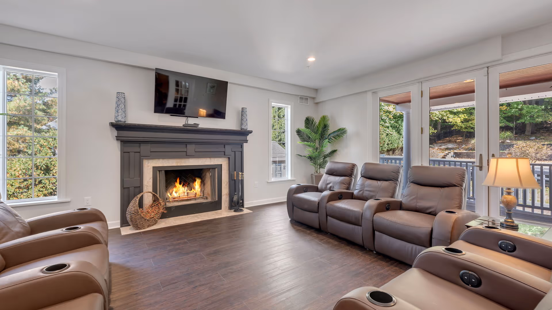 Bright communal living room with a row of leather recliners facing a lit fireplace and wall-mounted TV, with large windows and glass doors to the outside.