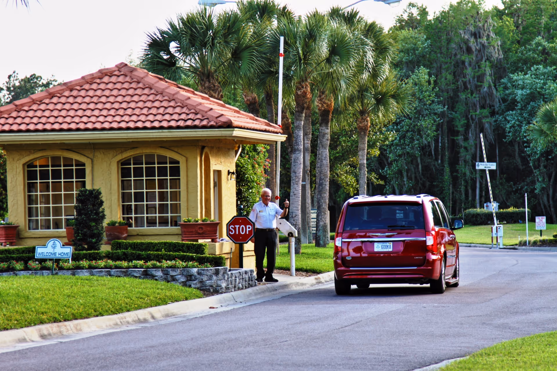 A red vehicle approaches a gated entrance with a small guardhouse featuring a red tiled roof and large windows. A security guard stands next to a stop sign, giving a thumbs-up. Palm trees and lush greenery surround the area, and a 'Welcome Home' sign is visible near the guardhouse.