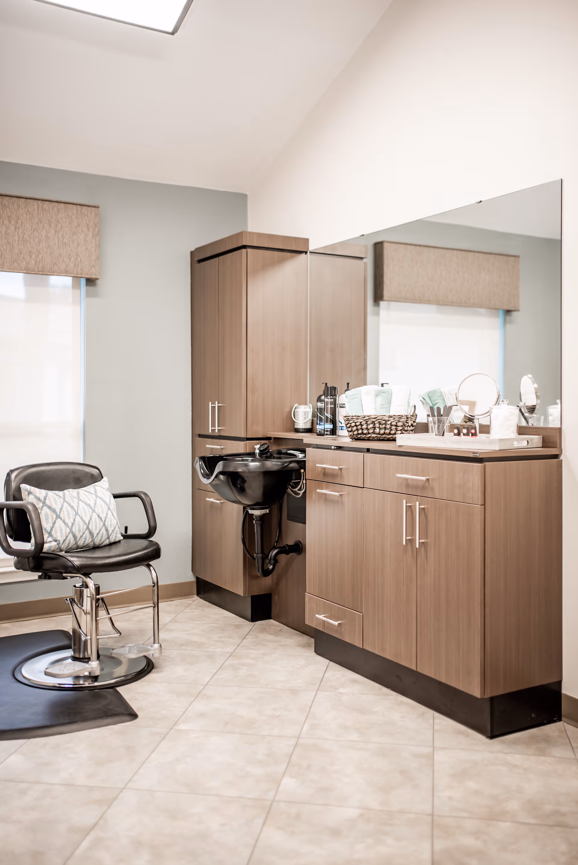 A clean and modern hair salon area inside a nursing and rehabilitation center, featuring a black salon chair with a patterned pillow, a black shampoo sink, wooden cabinets, a large mirror, and various hair care products neatly arranged on the countertop.