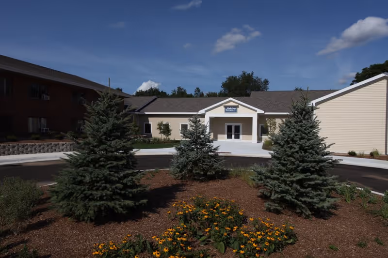 Exterior view of a senior living facility building with a beige facade and a peaked entrance. There are three evergreen trees and a flower bed with yellow flowers in the foreground under a partly cloudy blue sky.