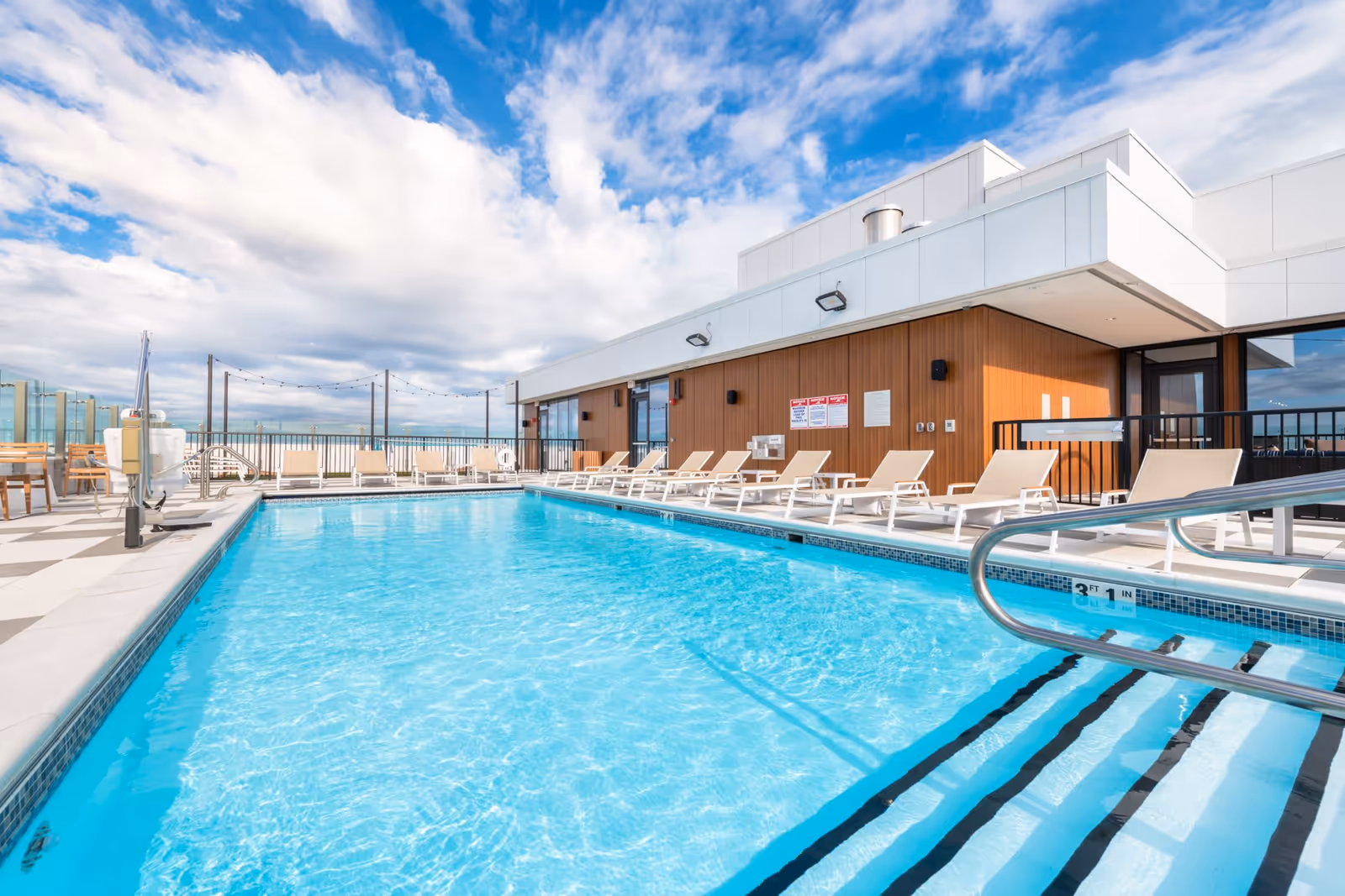Rooftop swimming pool with lounge chairs beside a modern building under a bright blue sky.
