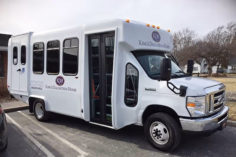 A white shuttle bus parked in a parking lot with the logo and name 'King's Daughters Home' on the side and front. The bus has large windows and a door in the middle, with a handicap accessibility symbol near the rear door. Trees and houses are visible in the background.