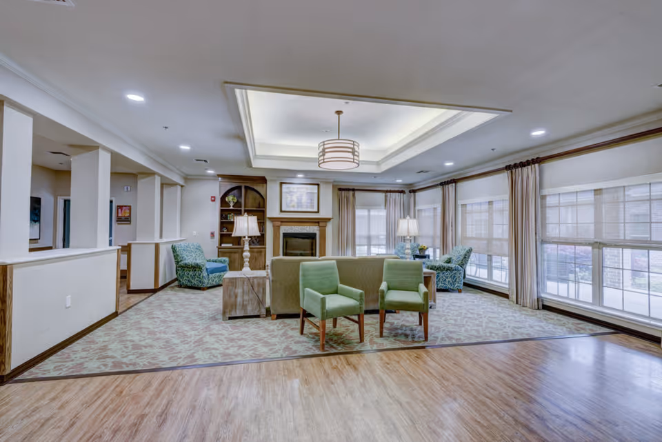 Bright communal living room with seating including two green chairs, a sofa, a fireplace, lamps, and large windows.