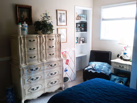 A cozy bedroom with a blue bedspread, a vintage-style dresser with multiple drawers, a small black chair with a blue and white blanket draped over it, a nightstand with a lamp and decorative items, framed pictures on the wall, and a window letting in natural light.