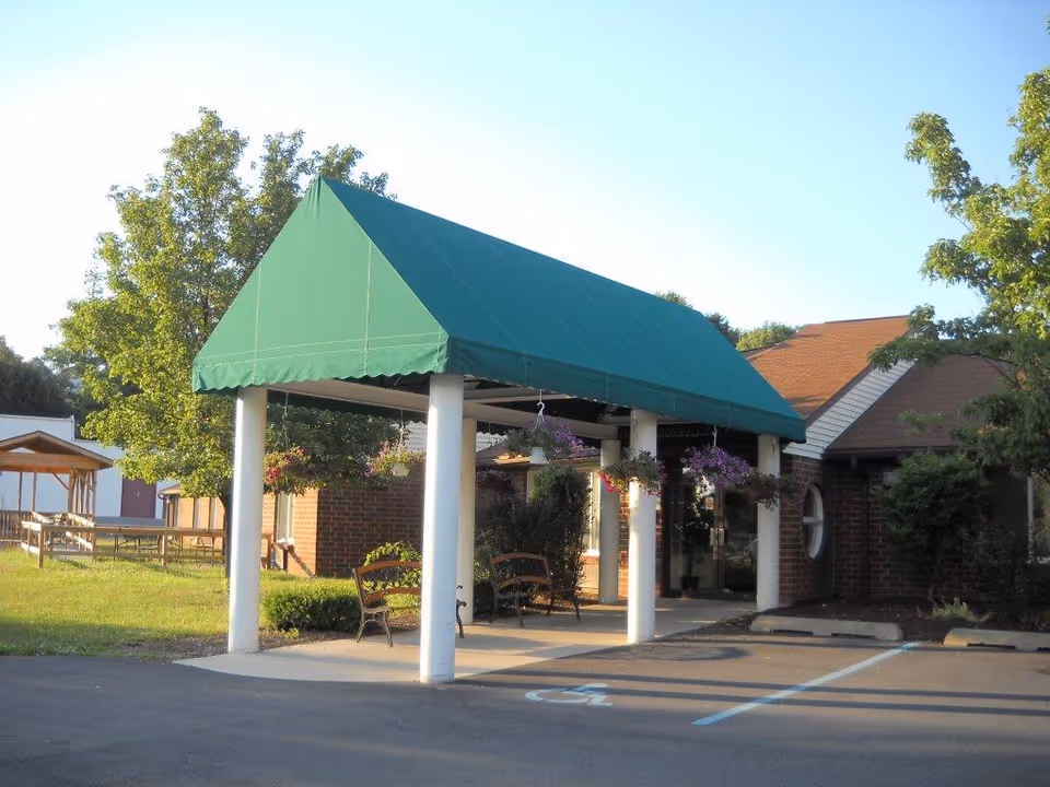 Entrance of a brick building with a green canopy supported by white columns. There are benches under the canopy and hanging flower baskets. Trees and a grassy area are visible around the building, along with a parking lot in the foreground.