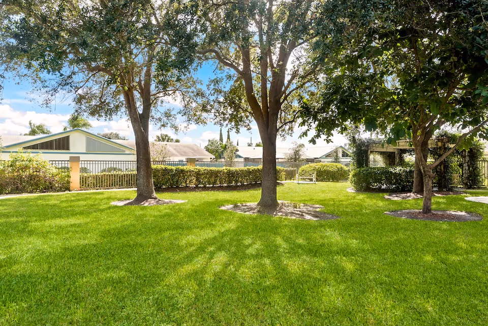 A well-maintained outdoor garden area with green grass, several trees providing shade, and a bench near a hedge and fence. Residential buildings are visible in the background under a partly cloudy sky.