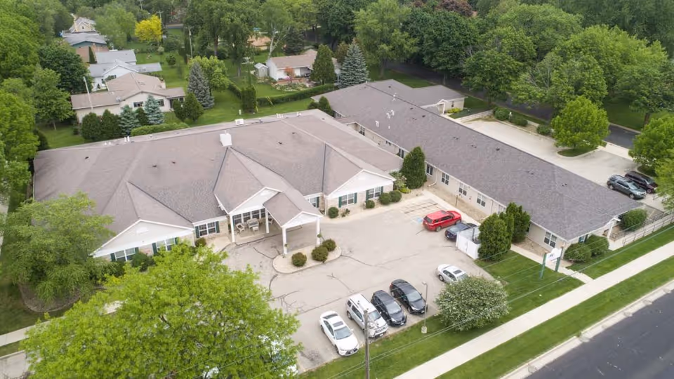 Aerial view of Madison Pointe Senior Living facility showing a large, single-story building with a gray roof surrounded by trees and greenery. There is a parking lot with several cars in front of the building and a sidewalk along the street.
