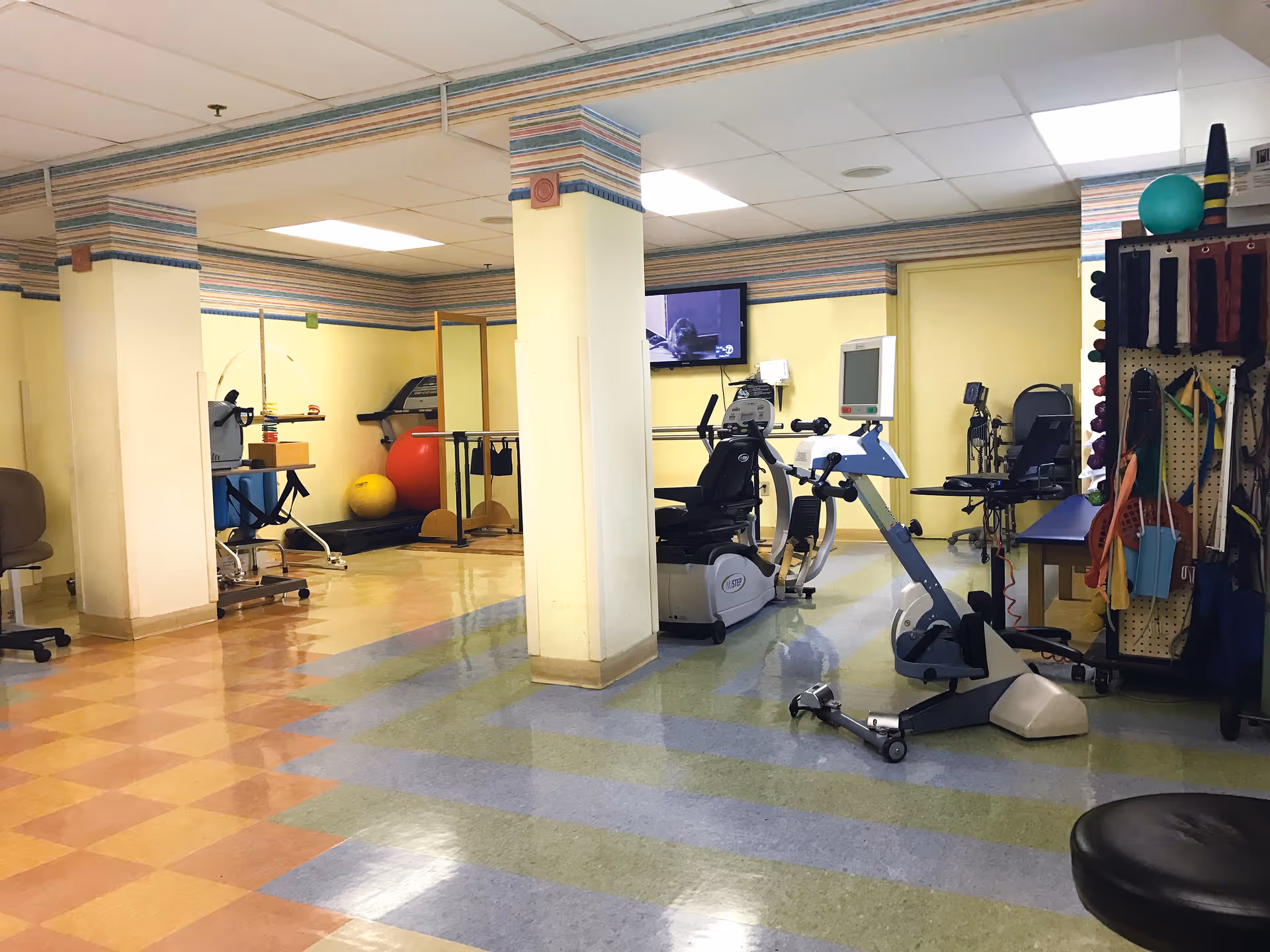 Interior view of a rehabilitation or physical therapy room with exercise equipment including a recumbent bike, treadmill, therapy balls, and various therapy tools hanging on a pegboard. The room has yellow walls with a striped border near the ceiling and a checkered floor with blue and green patterns.