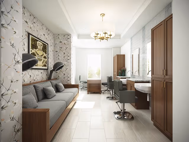 A bright, modern interior room with a gray sofa and cushions on the left side, floral patterned wallpaper behind it, two black hair dryer chairs mounted on the wall, and a framed artwork above the sofa. On the right side, there are two salon chairs in front of a counter with sinks and wooden cabinets. A window at the far end lets in natural light, and a chandelier hangs from the ceiling.