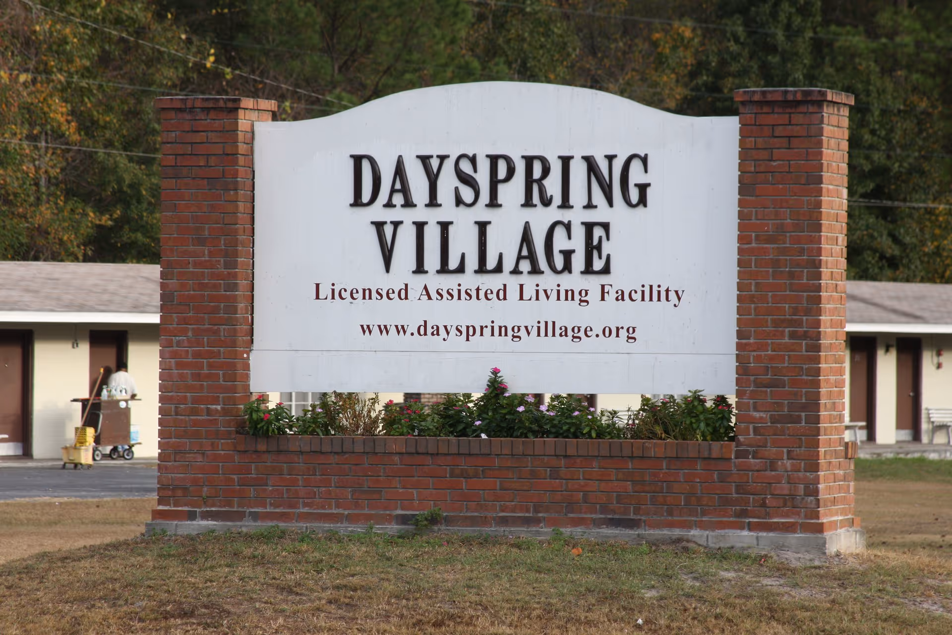 A large white sign with black and brown text reading 'DAYSPRING VILLAGE Licensed Assisted Living Facility www.dayspringvillage.org' mounted on a brick base with two brick pillars on each side, with some greenery and flowers planted in front. In the background, there are single-story buildings and a person pushing a cleaning cart.