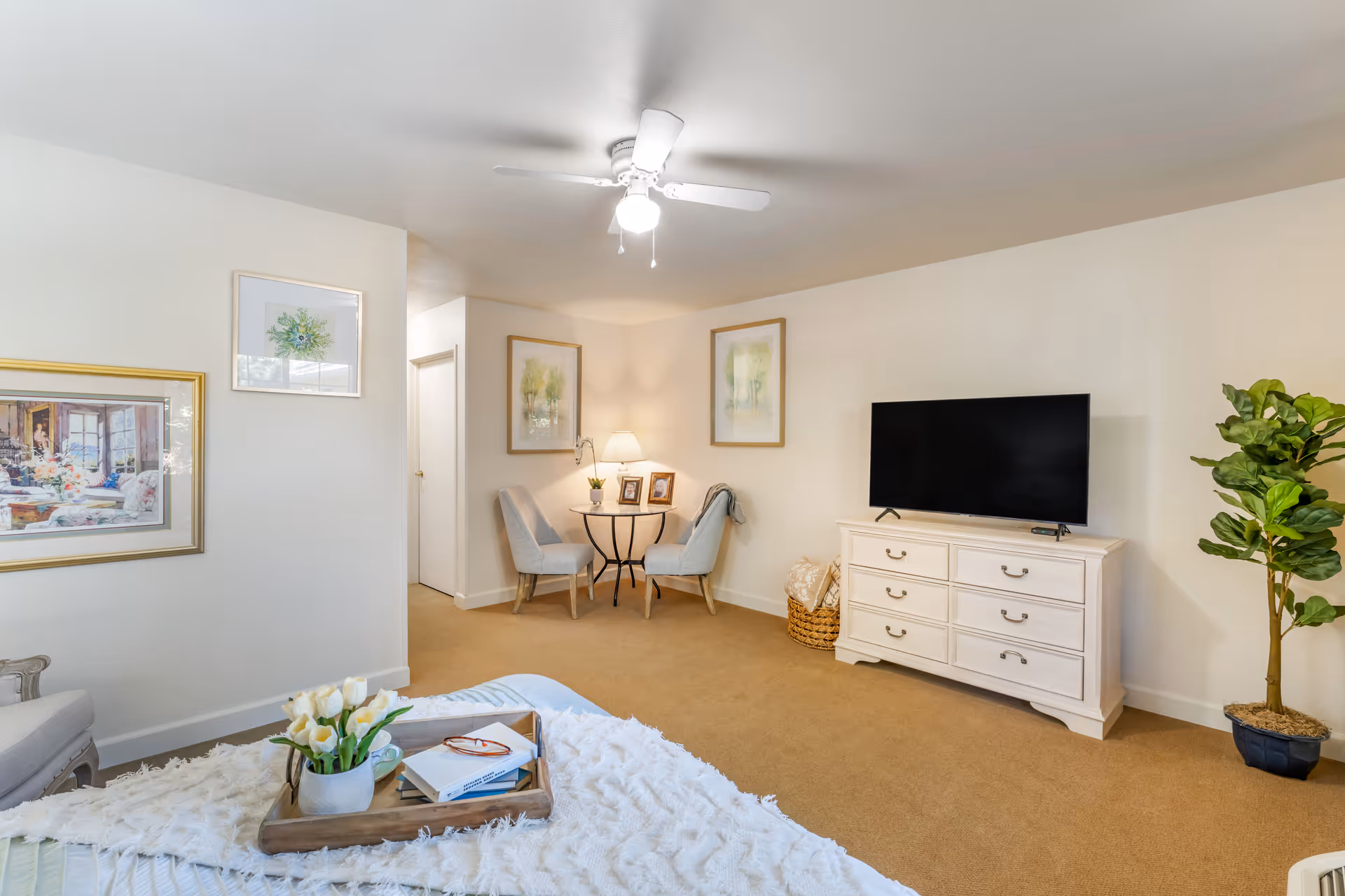 A cozy living room area with beige carpet and cream-colored walls. There is a white dresser with a flat-screen TV on top, a small round table with two upholstered chairs, framed artwork on the walls, a ceiling fan with a light, and a potted plant in the corner. A bed with a white textured blanket and a wooden tray holding a vase of white tulips, books, and glasses is partially visible in the foreground.