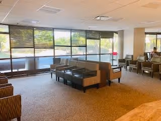 A spacious seating area in a senior living facility with multiple chairs and small tables arranged near large windows that let in natural light. The room has carpeted floors and a neutral color scheme, with a person sitting in the background near the windows.