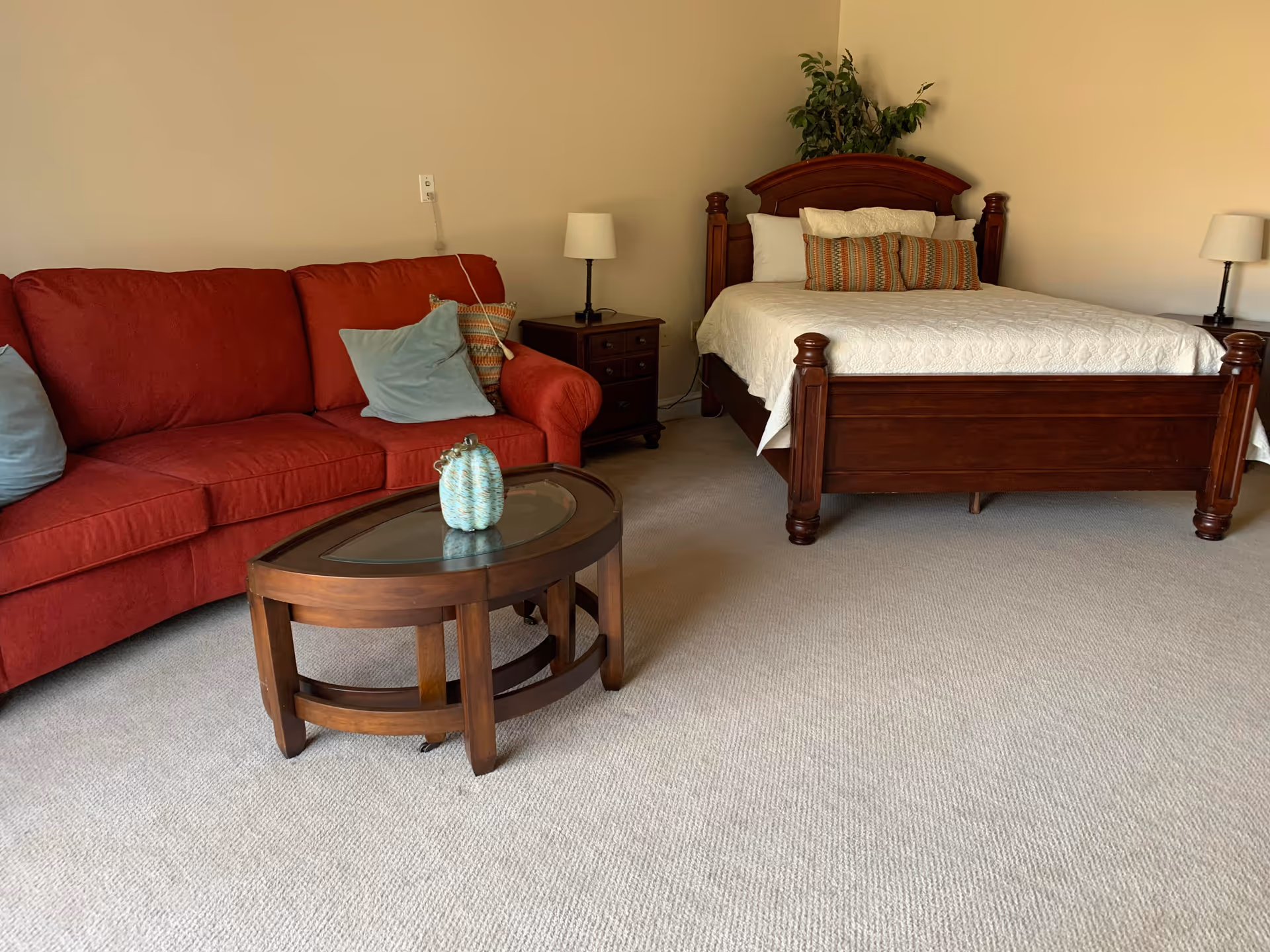 A cozy room featuring a wooden bed with white bedding and decorative pillows, a red sofa with blue and patterned cushions, a wooden coffee table with a small decorative item, two bedside tables each with a lamp, and a plant in the corner against beige walls and carpeted floor.