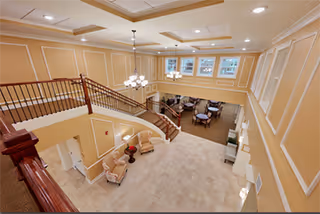 View from an upper level overlooking a spacious, elegant common area with a curved staircase, beige walls with white trim, tiled floor, and seating areas with chairs and tables near windows.