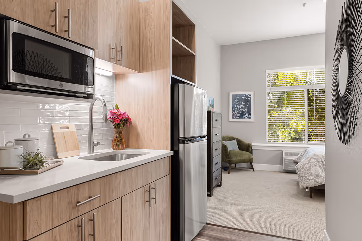 A modern studio apartment with a small kitchen area featuring light wood cabinets, a stainless steel microwave, sink, and refrigerator. The kitchen opens into a living and sleeping area with a green armchair, a dresser, a bed with patterned bedding, a window with white blinds, and a decorative round mirror on the wall.