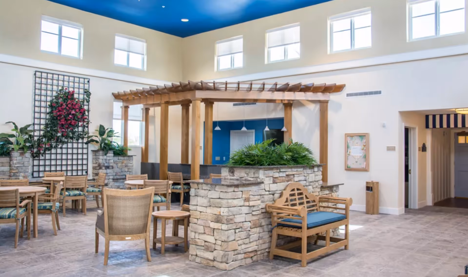 Bright and spacious indoor common area with high ceilings and multiple windows. The room features wooden chairs and tables with patterned cushions, a wooden bench with a blue cushion, and a stone planter with green plants. A wooden pergola structure is centrally located, and there is a decorative trellis with pink flowers on one wall.