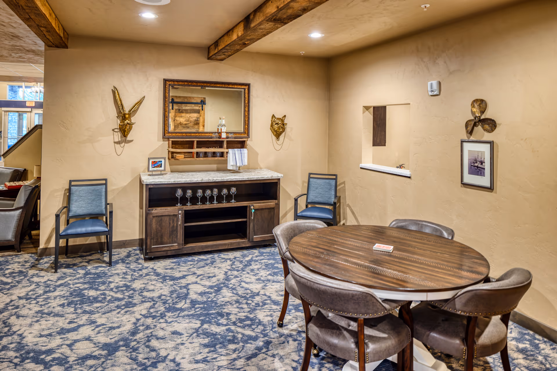 Cozy senior living common area with a round wooden table and chairs, a sideboard holding glassware, and decorative wall art on beige walls.
