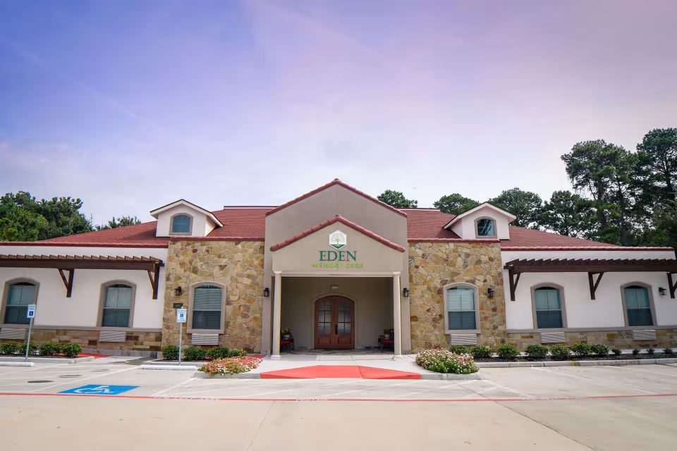 Front exterior view of a single-story memory care facility building with a red roof, stone and beige walls, and a covered entrance. The building has multiple windows and is surrounded by a parking lot with handicap parking spaces and landscaped flower beds. Trees are visible in the background under a purple-tinted sky.