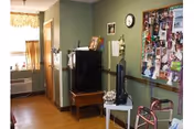 Interior view of a room in a senior living facility with green walls and wooden flooring. There is a small black refrigerator on a wooden table, a white table with a black fan and a telephone, a bulletin board filled with photos and notes, a clock on the wall, and a walker in the foreground. A window with curtains and an air conditioning unit is visible in the background.