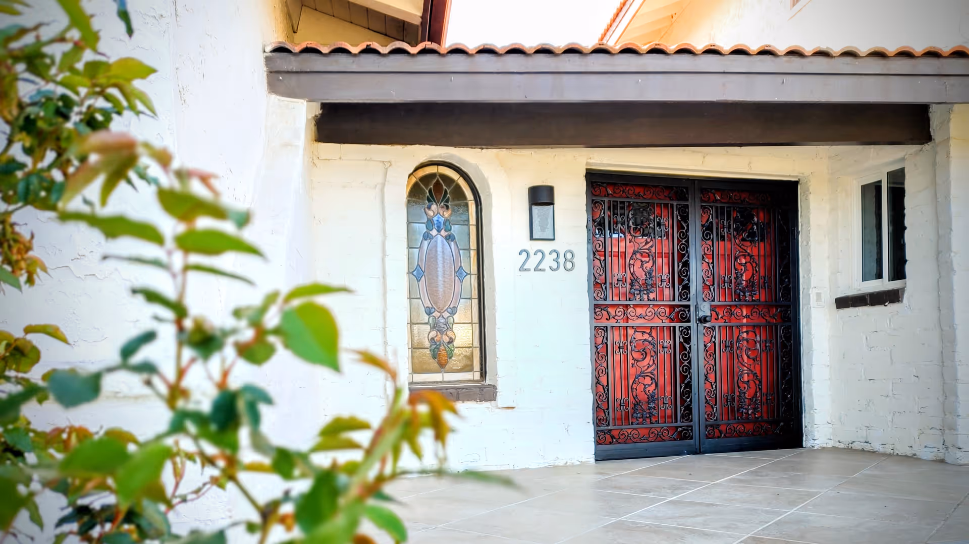 Front entrance of a building with white brick walls, a decorative stained glass window, the number 2238 displayed next to a black wall-mounted light fixture, and a double door with intricate black wrought iron designs over red panels. Some green foliage is visible in the foreground.