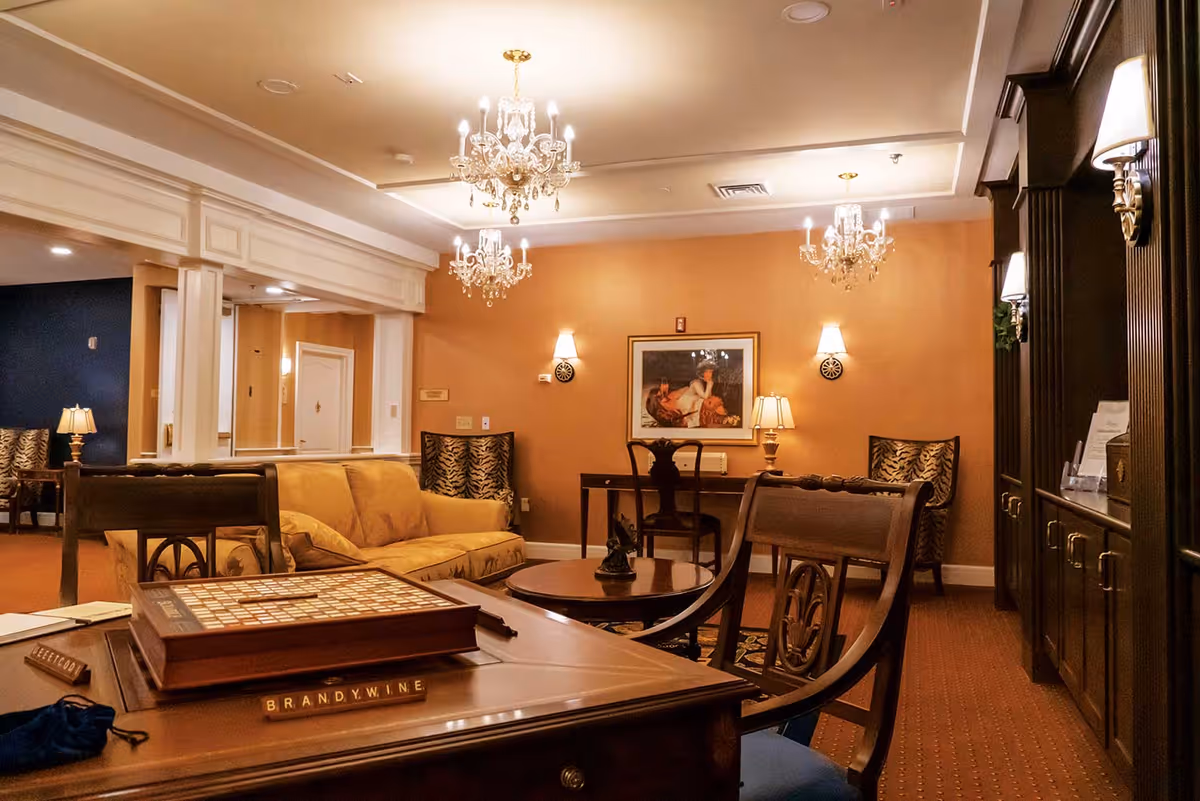 A warm, elegantly furnished senior living lounge with chandeliers, sofas, chairs, and a wooden game table.