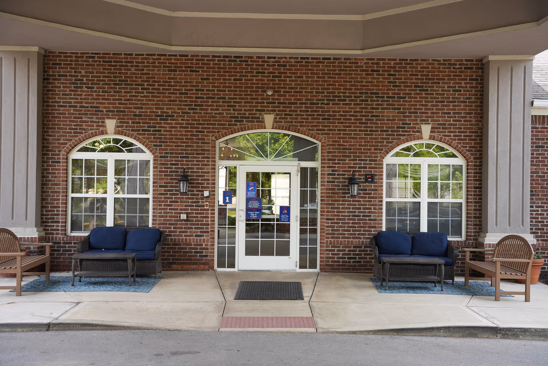 Front entrance of a brick building with double glass doors flanked by arched windows and outdoor seating with blue-cushioned benches.