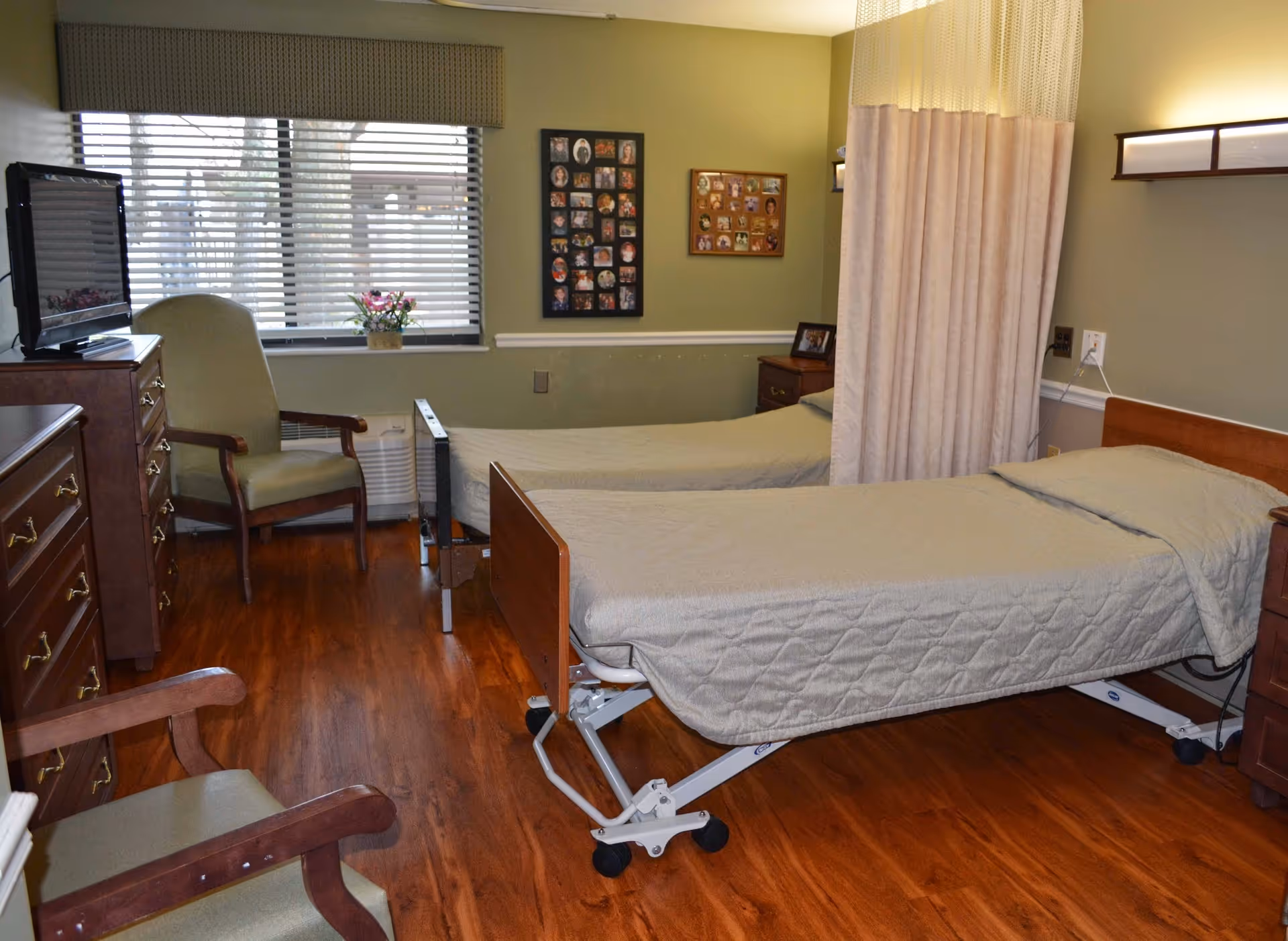 A nursing facility room with two hospital-style beds covered in beige quilts, separated by a privacy curtain. The room has wooden flooring, two green armchairs, a wooden dresser with a TV on top, and two photo collages on the wall. A window with blinds lets in natural light.