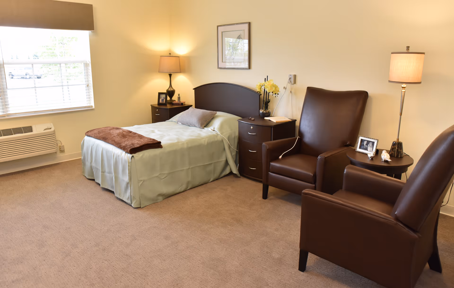 A neatly arranged bedroom in a senior living facility with a single bed covered in light green bedding and a brown throw blanket. There are two dark brown leather armchairs with a small round table between them, holding a lamp and framed photos. The room has beige carpet, a window with blinds, and soft lighting from two lamps.